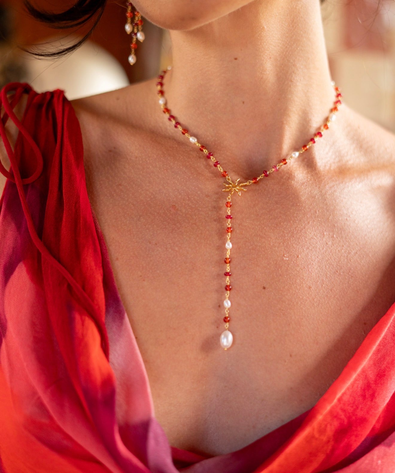 A close-up lifestyle shot of a woman wearing a delicate gold Y-necklace with red beads and white pearl accents, paired with a red and pink draped silk scarf.