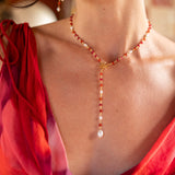 A close-up lifestyle shot of a woman wearing a delicate gold Y-necklace with red beads and white pearl accents, paired with a red and pink draped silk scarf.