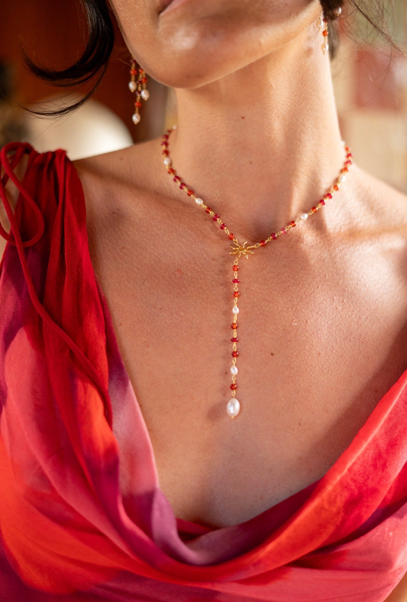A close-up lifestyle shot of a woman wearing a delicate gold Y-necklace with red beads and white pearl accents, paired with a red and pink draped silk scarf.