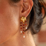 A close-up of a woman's ear wearing a dangling gold earring with red and white beads, against a softly blurred warm-toned background.