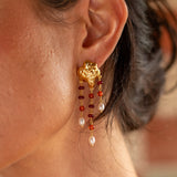 A close-up of a woman's ear wearing a dangling gold earring with red and white beads, against a softly blurred warm-toned background.
