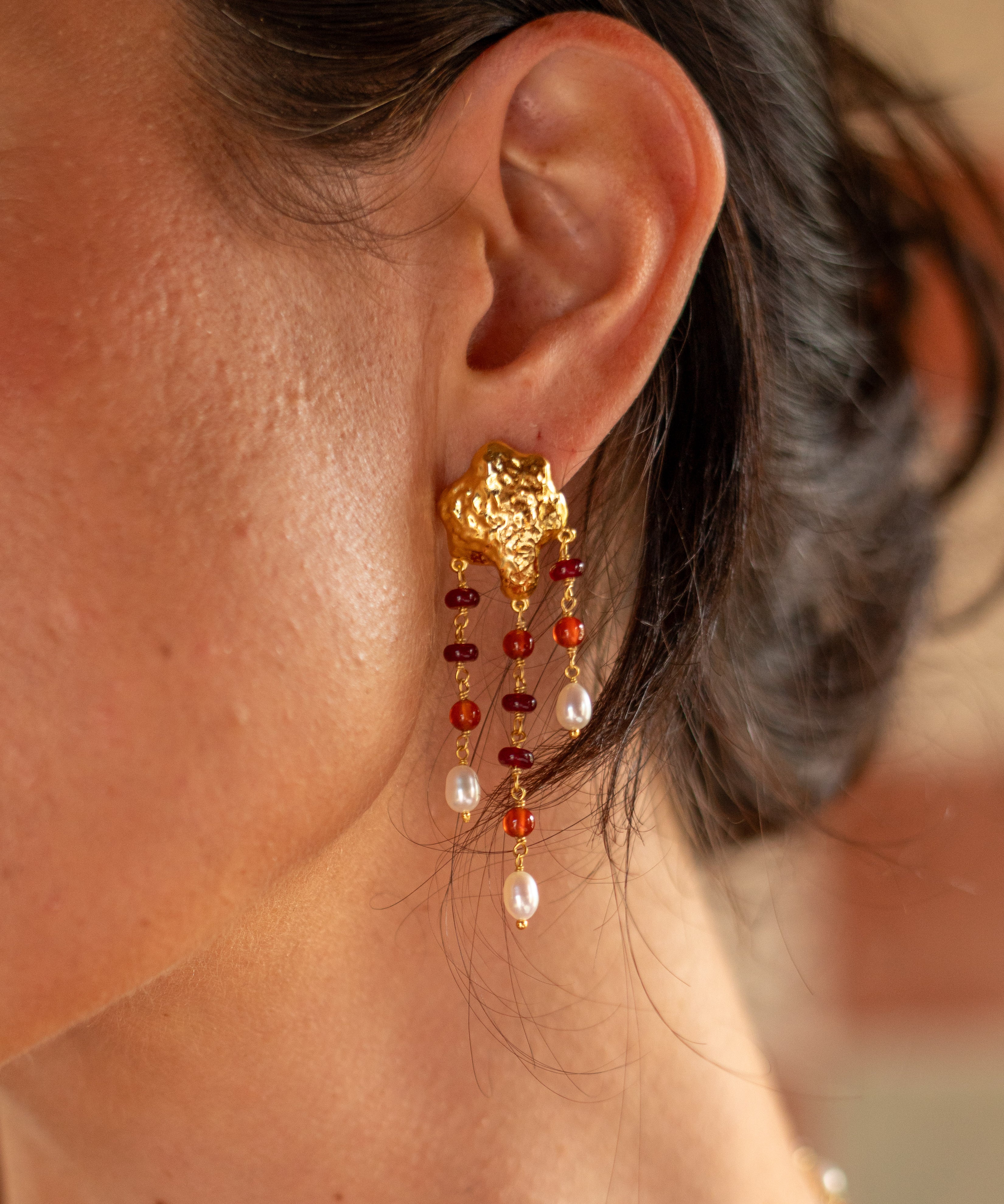 A close-up of a woman's ear wearing a dangling gold earring with red and white beads, against a softly blurred warm-toned background.