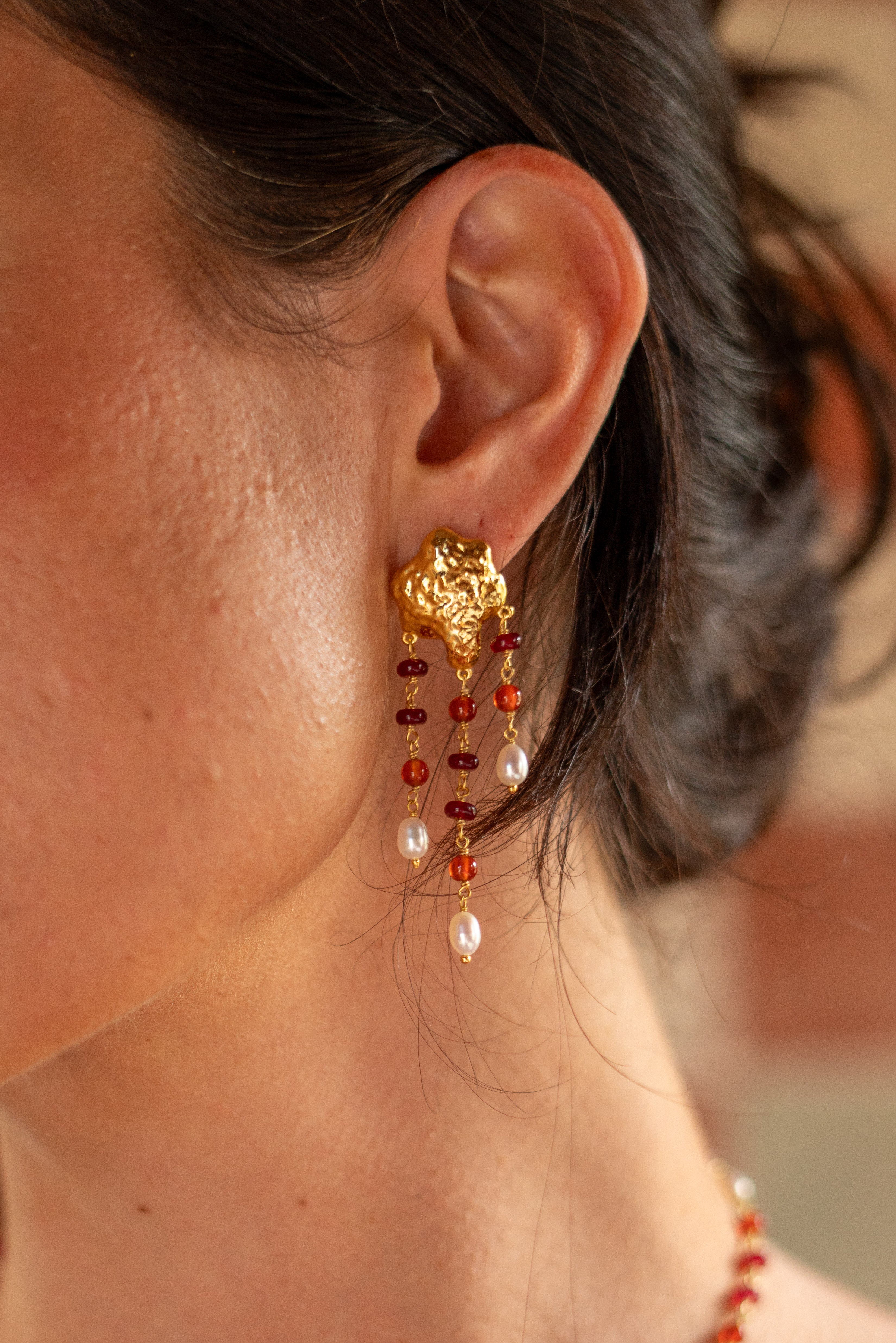 A close-up of a woman's ear wearing a dangling gold earring with red and white beads, against a softly blurred warm-toned background.
